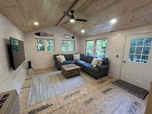 Une pièce avec un plafond en bois, un ventilateur de plafond, un canapé gris agrémenté de coussins à motifs, une table basse en bois et une grande fenêtre.