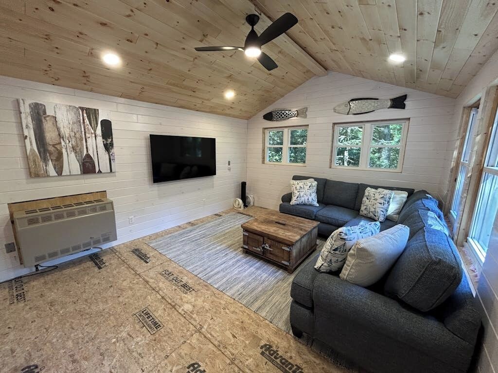 A living room with a wooden ceiling, a flat-screen TV, a gray sofa, and a window with a view of trees.