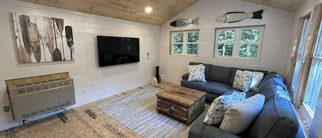 A living room with a wooden ceiling, a flat-screen TV, a gray sofa, and a window with a view of trees.