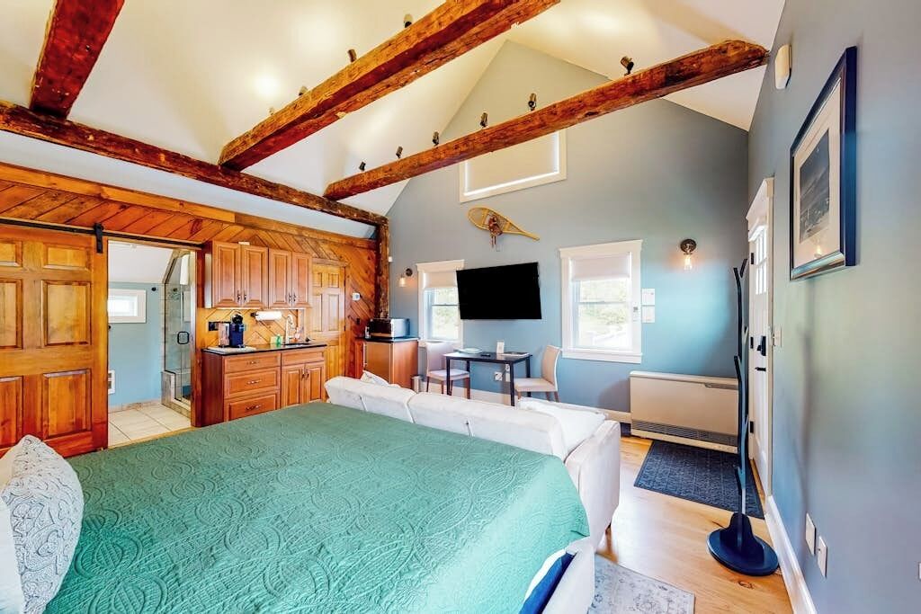 Une chambre avec un grand lit, des poutres apparentes, un coin cuisine, une télévision et une vue sur l’extérieur.