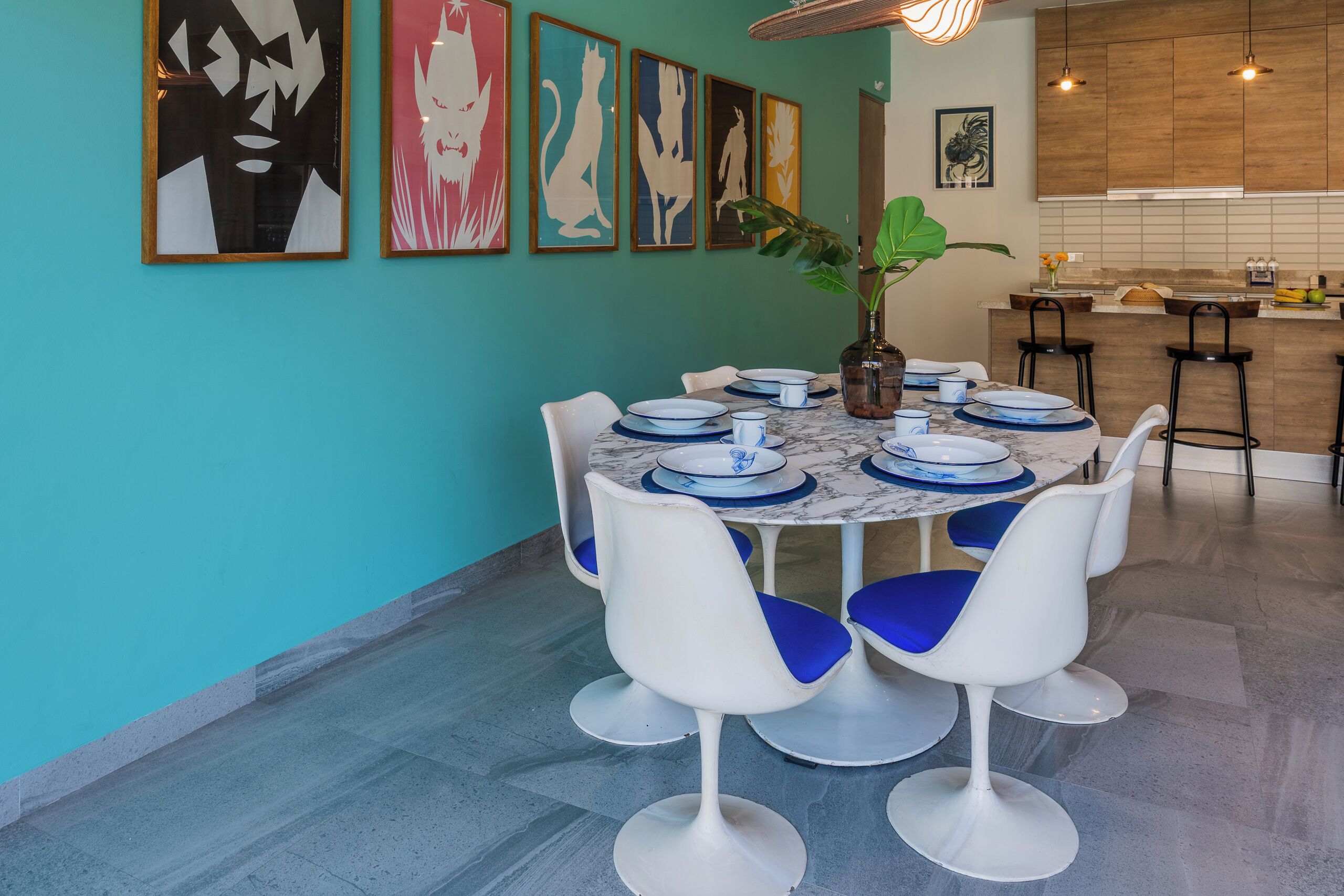 Un coin repas avec une table ronde, des chaises blanches et des coussins bleus. Des œuvres d’art encadrées ornent le mur, ainsi qu’une plante en pot. On aperçoit également une cuisine au fond.