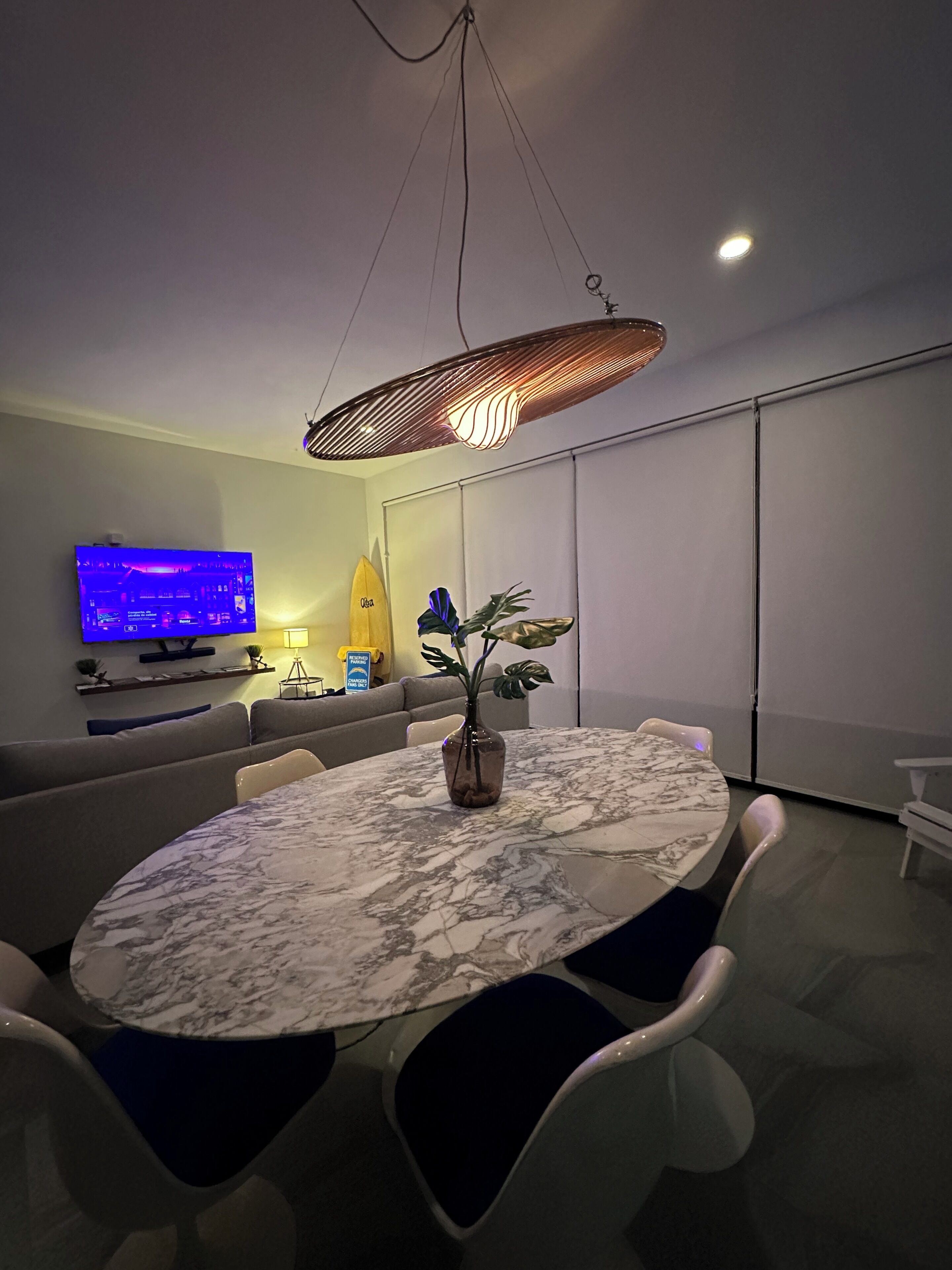 Ein moderner Essbereich mit einem Marmortisch, weißen Stühlen, einer Vase mit Blumen, einem Fernseher und einem Sofa.