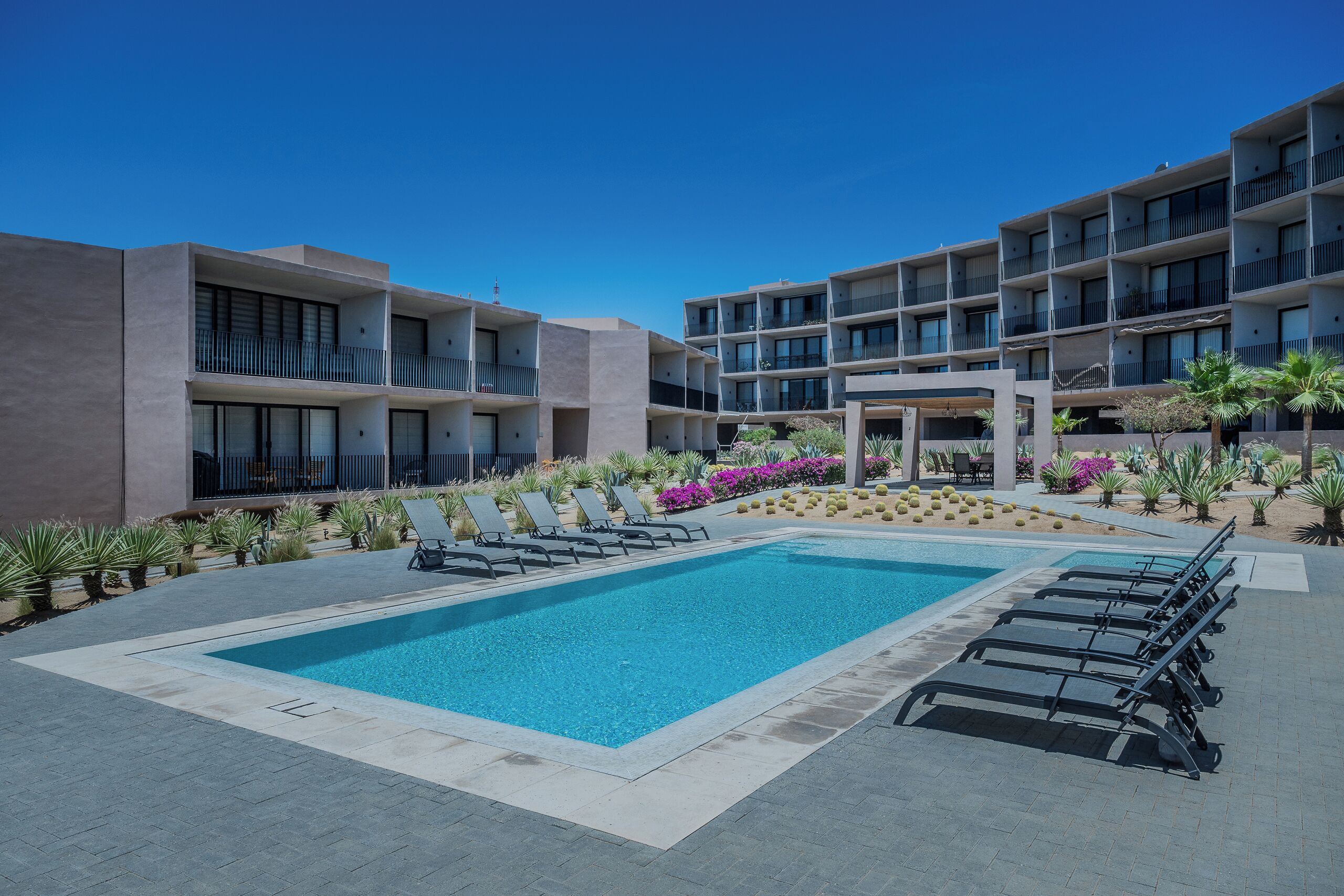 A swimming pool with lounge chairs, a covered seating area, and a building with balconies.