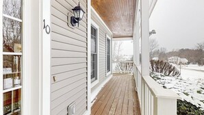 A covered porch with a wooden floor, a wall-mounted lantern, and a view of a snowy landscape. - Cozy home near park, deck, patio, bbq, soaking tub, TV, basketball, office, w/d (South Burlington)