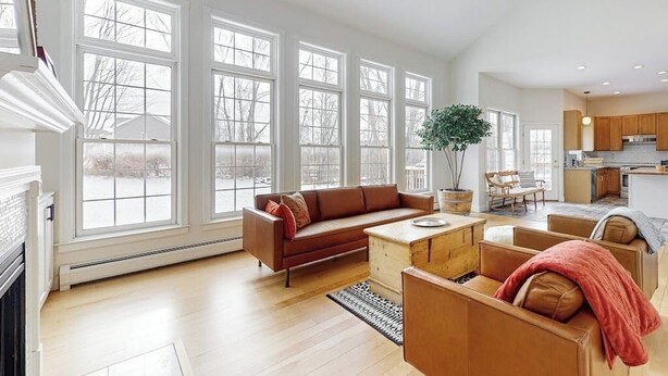 A spacious living area with large windows, a fireplace, and a kitchen in the background. - Cozy home near park, deck, patio, bbq, soaking tub, TV, basketball, office, w/d (South Burlington)