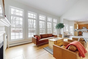 A spacious living area with large windows, a fireplace, and a kitchen in the background.