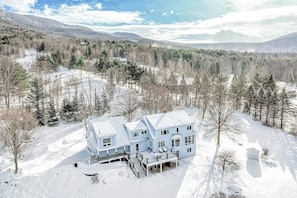 A large, light blue house with a covered porch and a staircase leading to the entrance, surrounded by snow-covered trees and a mountainous backdrop.