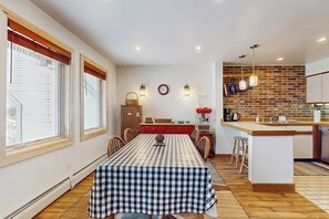 Un comedor con mantel a cuadros, una cocina con salpicadero de ladrillo y un reloj en la pared.