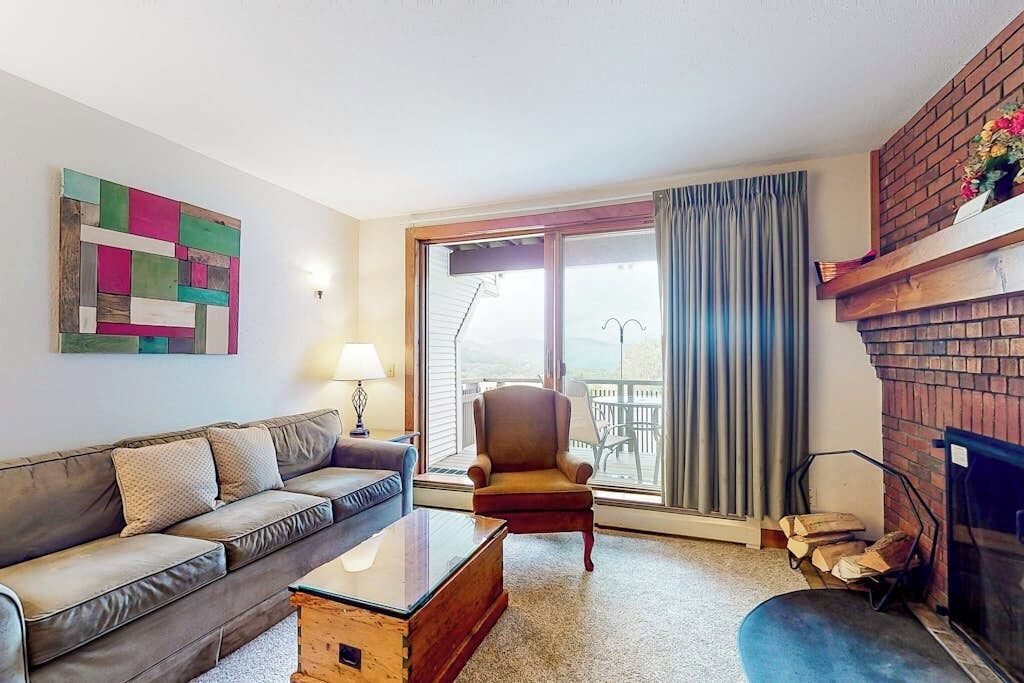 Un salon comprenant un canapé, un fauteuil, une cheminée et une vue dégagée sur l’extérieur grâce à une grande fenêtre.