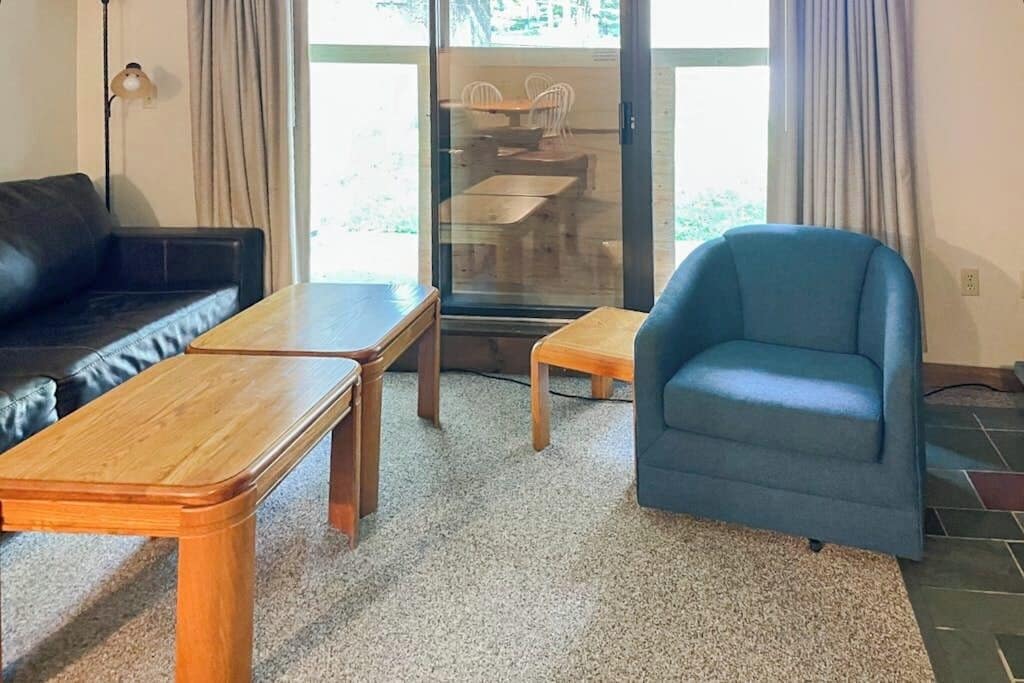 Una sala de estar con un sofá, dos mesas de madera, un sillón azul y un ventanal de vidrio que da a un comedor al aire libre.