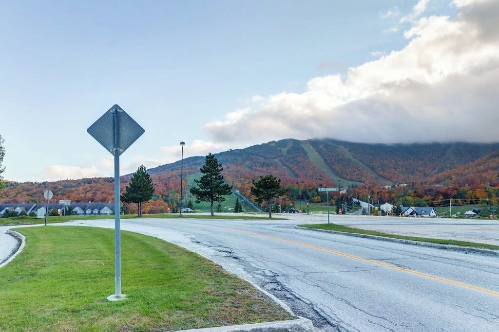 A road with a diamond-shaped sign, a grassy area, and a mountain with autumn foliage in the background.