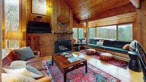 A cozy cabin interior with a wooden interior, a fireplace, a sofa, and a television. - Ski home in the woods with a private hot tub & mountain views (Fayston)
