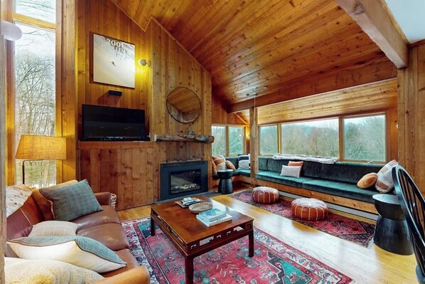 A cozy cabin interior with a wooden interior, a fireplace, a sofa, and a television. - Ski home in the woods with a private hot tub & mountain views (Fayston)
