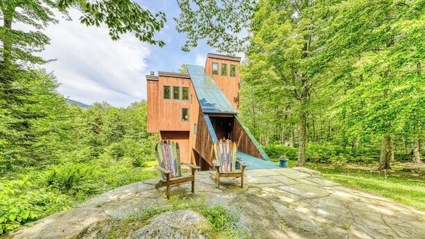 A modern building with a unique sloped roof, surrounded by lush greenery and two wooden chairs. - Ski home in the woods with a private hot tub & mountain views (Fayston)