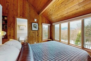 A wooden cabin bedroom with a large bed, a window with a view of trees, and a wooden ceiling. - Ski home in the woods with a private hot tub & mountain views (Fayston)