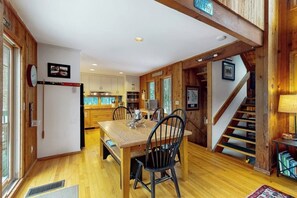 A wooden interior with a dining table, chairs, a kitchen area, and a staircase. - Ski home in the woods with a private hot tub & mountain views (Fayston)