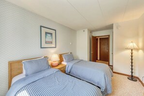 Una habitación con dos camas, una mesita de noche, una lámpara, un cuadro en la pared y una puerta que da a otra habitación.