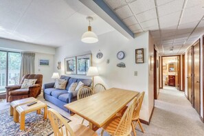 Ein Wohnzimmer mit einer Couch, einem Sessel, einem Holztisch und Stühlen. Ein Fenster mit Vorhängen, eine Lampe und Wanddekoration sind vorhanden.