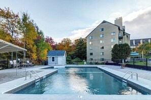 Una piscina con un edificio pequeño adyacente, rodeada de árboles con follaje otoñal, y un edificio de varios pisos al fondo.