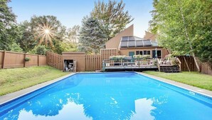 Ein Schwimmbad mit Holzterrasse, einer überdachten Terrasse und einem Haus mit Solaranlage.