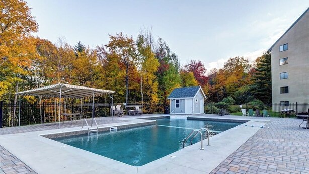 Una piscina rectangular con un área de estar techada y un edificio pequeño, rodeada de árboles con follaje otoñal.