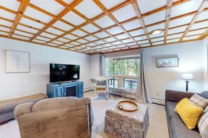 Ein Wohnzimmer mit Holzbalkendecke, einem Fernseher, einem blauen Schrank, einem grauen Sofa, einem gemusterten Hocker und einem Fenster mit Vorhängen.