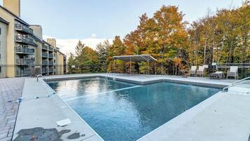 Una piscina rectangular con solárium metálico y una zona de estar cubierta, rodeada de árboles y un edificio con balcones.
