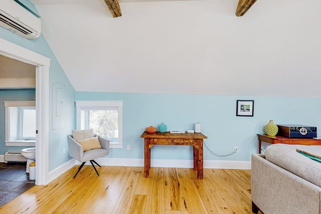 A room with a wooden floor, a grey armchair, a wooden table with decorative items, a white wall, and a window.