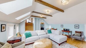 Uma sala de estar ampla com um sofá seccional grande, um banquinho redondo e uma mesa de centro de madeira. Há uma escada que leva a um andar superior, e o quarto tem um teto inclinado com vigas aparentes.