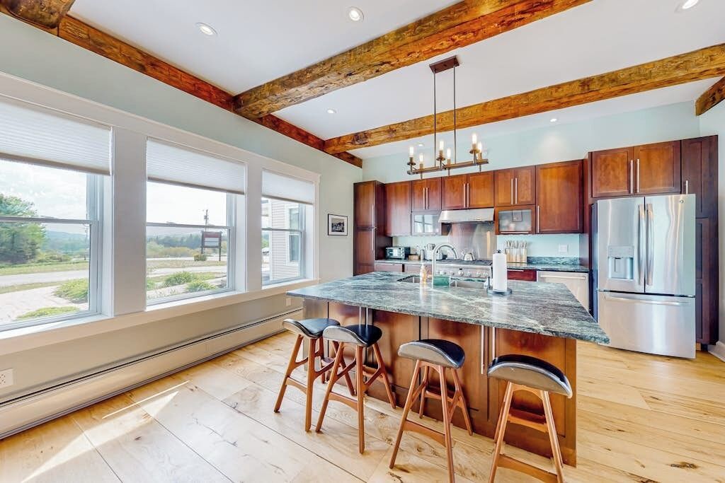 A modern kitchen with wooden beams, large windows, and a central island with stools.