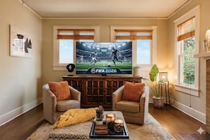 Sala de estar com uma grande TV exibindo uma partida da Copa do Mundo FIFA 2026, dois sofás de couro com almofadas laranja, uma mesa de centro com velas e uma planta.