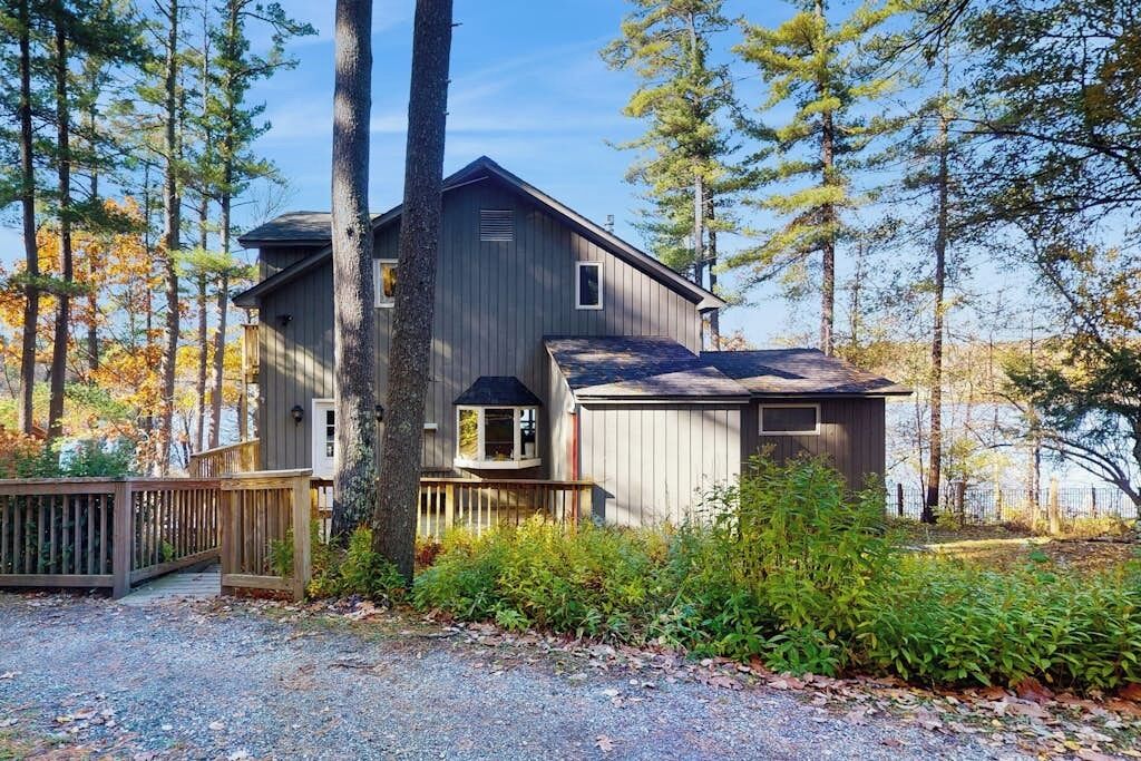 Une cabane à deux étages avec une terrasse, entourée d’arbres et donnant sur une allée en gravier.
