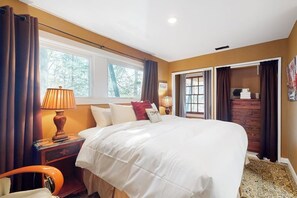 Ein Schlafzimmer mit einem großen Bett, einem Nachttisch mit Lampe, einer Holzkommode und einem Fenster mit Vorhängen.