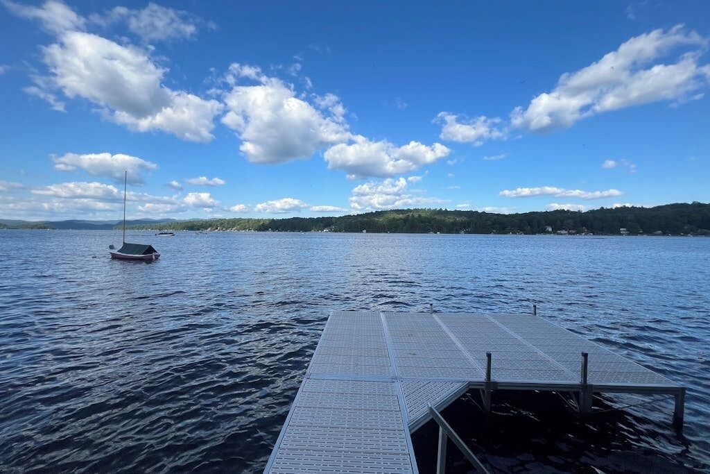 A calm lake with a dock extending into the water, a sailboat in the distance, and a hilly landscape under a partly cloudy sky.