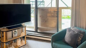 Un salon avec un téléviseur à écran plat posé sur un meuble en bois, un canapé turquoise agrémenté d’un coussin et une grande fenêtre donnant sur un paysage verdoyant.
