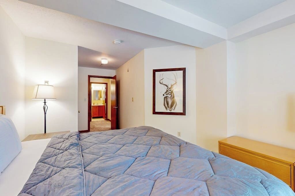 A hotel room with a bed, a bedside lamp, a wooden nightstand, a framed picture on the wall, and a door leading to another room.