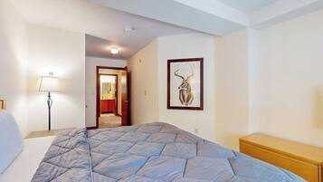 A hotel room with a bed, a bedside lamp, a wooden nightstand, a framed picture on the wall, and a door leading to another room.
