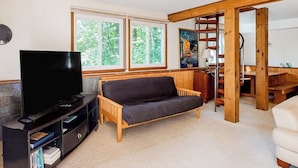 Ein Wohnzimmer mit einer Couch, einem Fernseher und einem Bücherregal. Fenster bieten Blick auf Bäume.