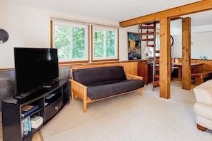 Ein Wohnzimmer mit einer Couch, einem Fernseher und einem Bücherregal. Fenster bieten Blick auf Bäume.