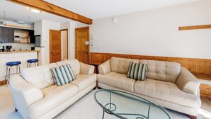 Ein Wohnzimmer mit einem beige gefärbten Sofa, zwei gestreiften Kissen, einem Glastisch und einem Holzbalken an der Decke.
