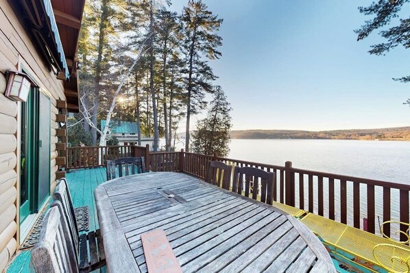 Eine Holzterrasse mit Tisch und Stühlen, die auf einen See mit Bäumen und Hügeln im Hintergrund blickt.