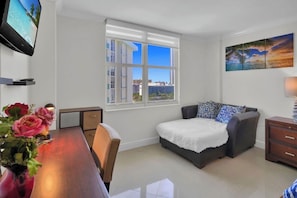 Ein modernes Zimmer mit Bett, Schreibtisch, Fernseher, Fenster mit Stadtblick und einem Bild an der Wand.