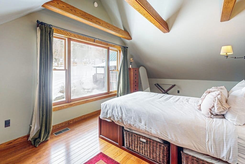 A bedroom with a large window, wooden beams, a bed, a lamp, and a dresser.