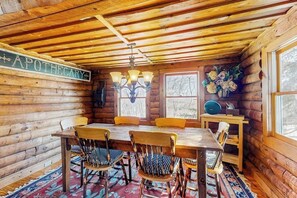 Interior de una cabaña de troncos con una mesa de comedor, sillas, una araña y un letrero que dice 'APOTHECARY'.