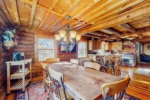 A rustic wooden cabin interior with a dining table, chairs, a chandelier, a kitchen area, and a large window. - Cozy log cabin & loft, close to recreation, dog-friendly, bbq, balcony, a/c, w/d (Barnard)
