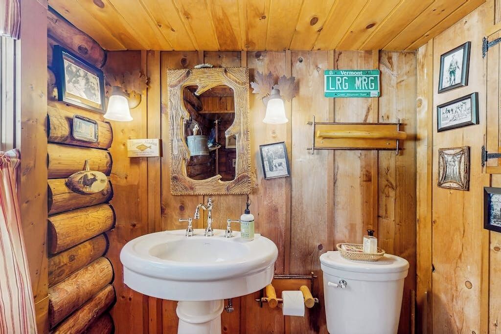 A rustic bathroom with a wooden log wall, a sink, a toilet, and framed pictures.