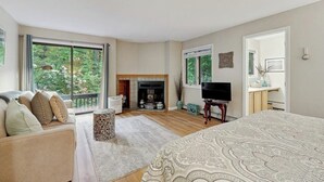 A bedroom with a large bed, a TV, a fireplace, and a balcony with a view of greenery.