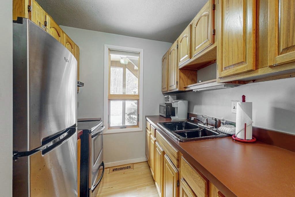 A kitchen with wooden cabinets, a stainless steel refrigerator, a microwave, a sink, and a window.