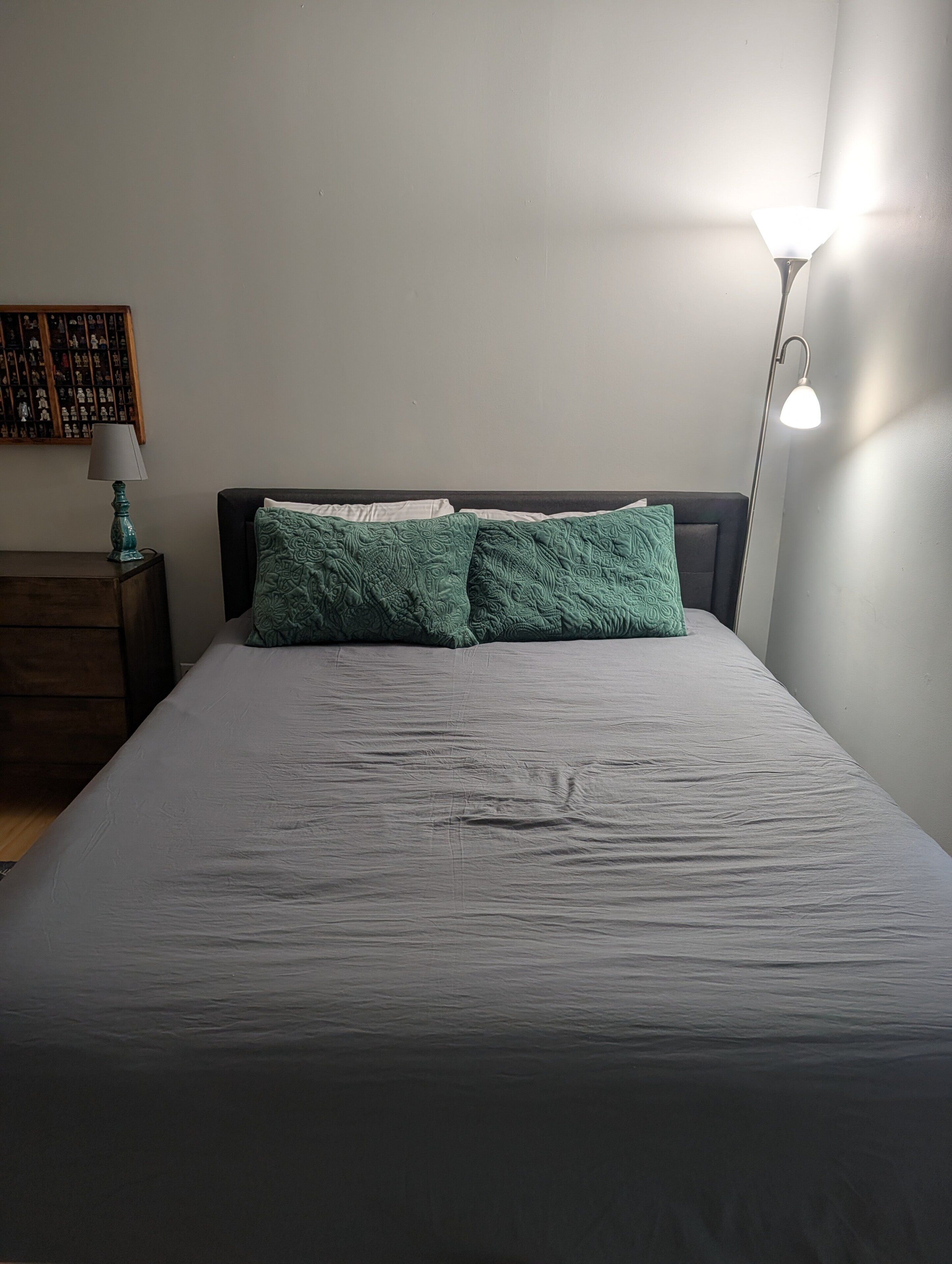 Ein Bett mit grauer Tagesdecke und zwei grünen Kissen, ein Nachttisch mit Lampe sowie eine Wand mit einem Regal voller Figuren.