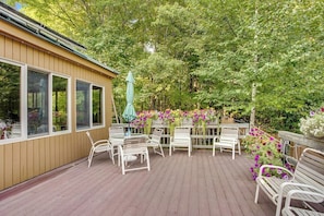 Eine Holzterrasse mit weißen Metallstühlen und Tischen, umgeben von üppiger Bepflanzung und blühenden Pflanzen.